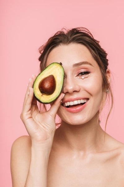 young woman smiling and holding an avocado in front of her right eye - pureyeva skincare