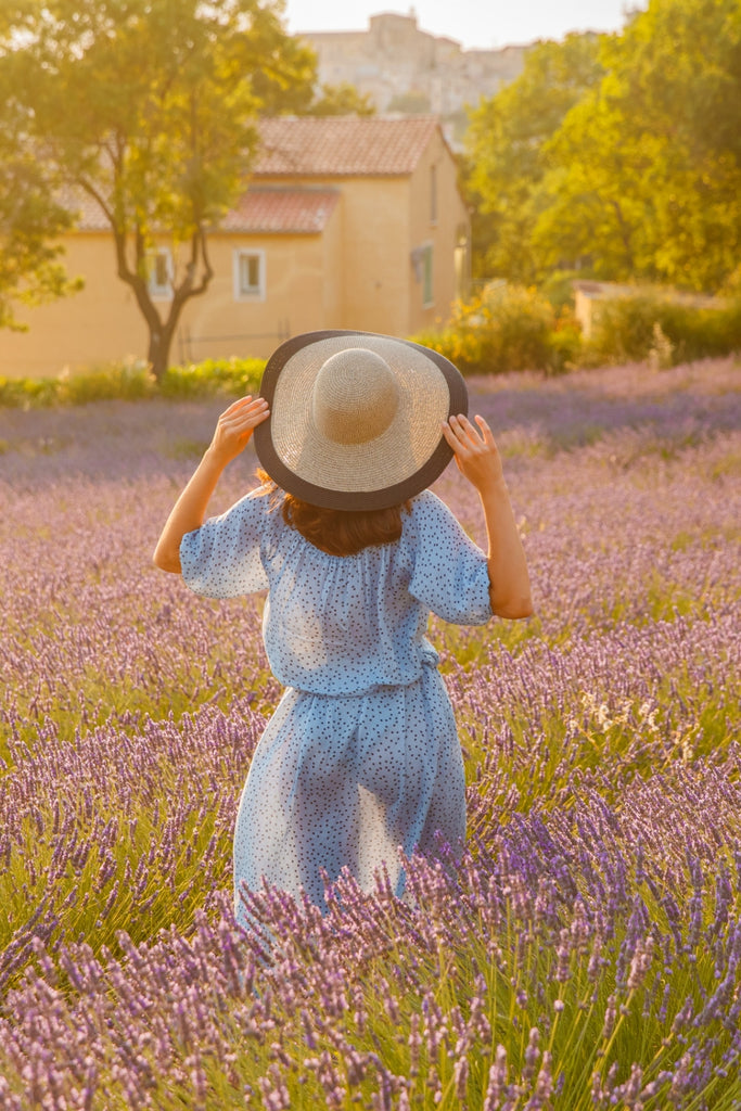 Woman with her back to us in a blue dress and cap walks across the field of a lavender flowers - pureyeva natural skincare 