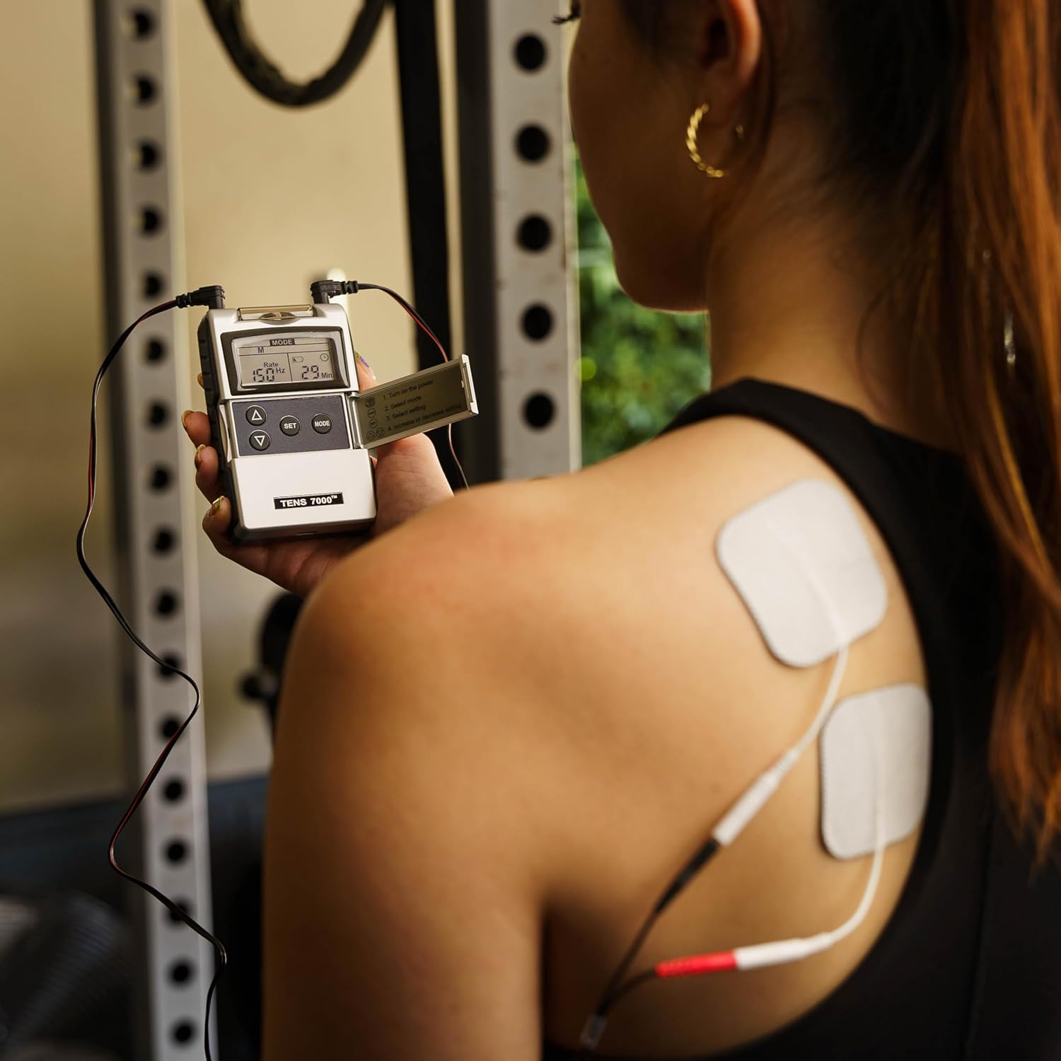 A gray TENS and EMS combo unit being held by a woman with electrodes on her back