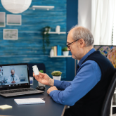 An elderly man holding up pills while speaking with his doctor over his computer