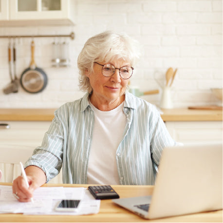An elderly woman sitting at her kitchen table with a calculator and laptop