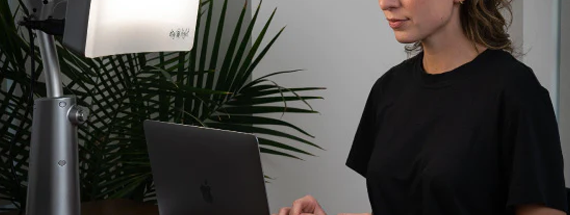 A woman working on a laptop at a desk with a bright light therapy lamp shining toward her