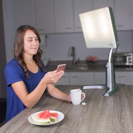 A woman on her phone sitting next to the Day-Light Classic Plus therapy lamp