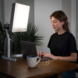 A woman working on her laptop basking under the Day-Light Classic Plus light therapy lamp