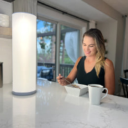 A woman eating breakfast next to the 24” Bright Health therapy lamp