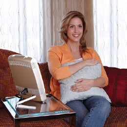 A woman sitting on a couch holding a pillow in front of the Carex Sunlite therapy lamp