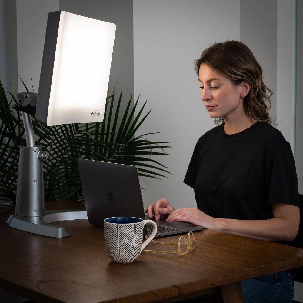 Woman sitting at a desk in front of the Day-Light Classic Plus light therapy lamp while working on a computer
