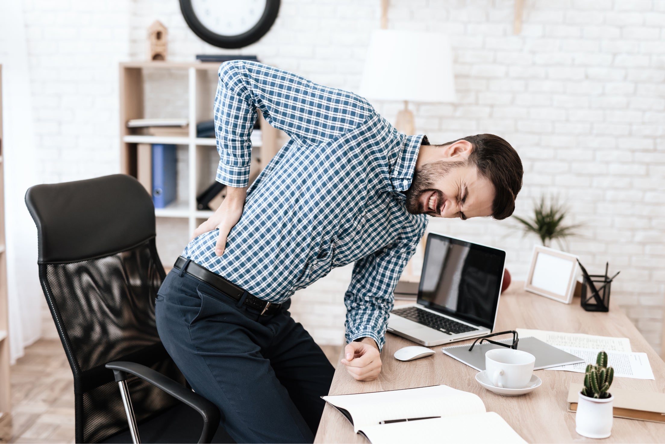 A man standing up from his desk holding his lower back in pain