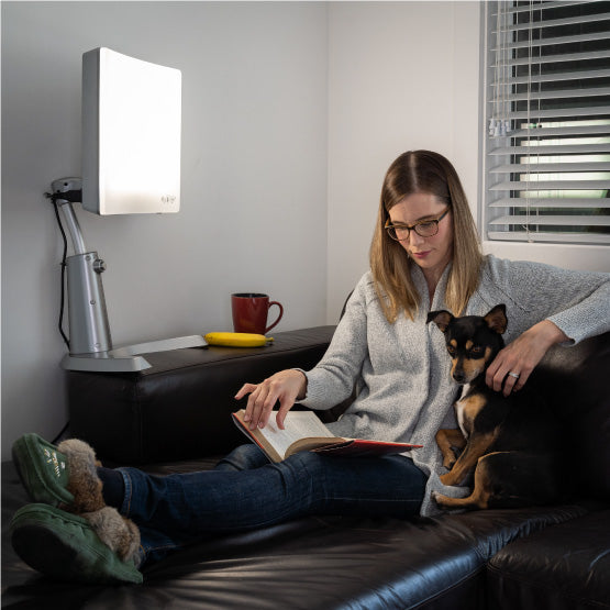 A woman sits on a couch with her dog, reading a book, and basking under the Day-Light Classic Plus light therapy lamp.