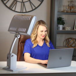 A woman at a desk in front of a computer in front of the Day-Light Sky therapy lamp.