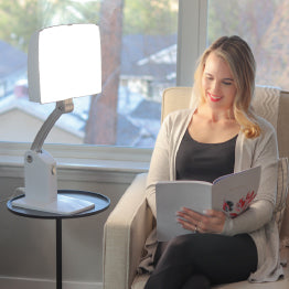 A woman sitting on a chair reading a book in front of the Day-Light Sky therapy lamp.