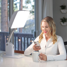 Woman sitting at a kitchen counter with a cup of coffee while basking in front of the Day-Light Sky light therapy lamp