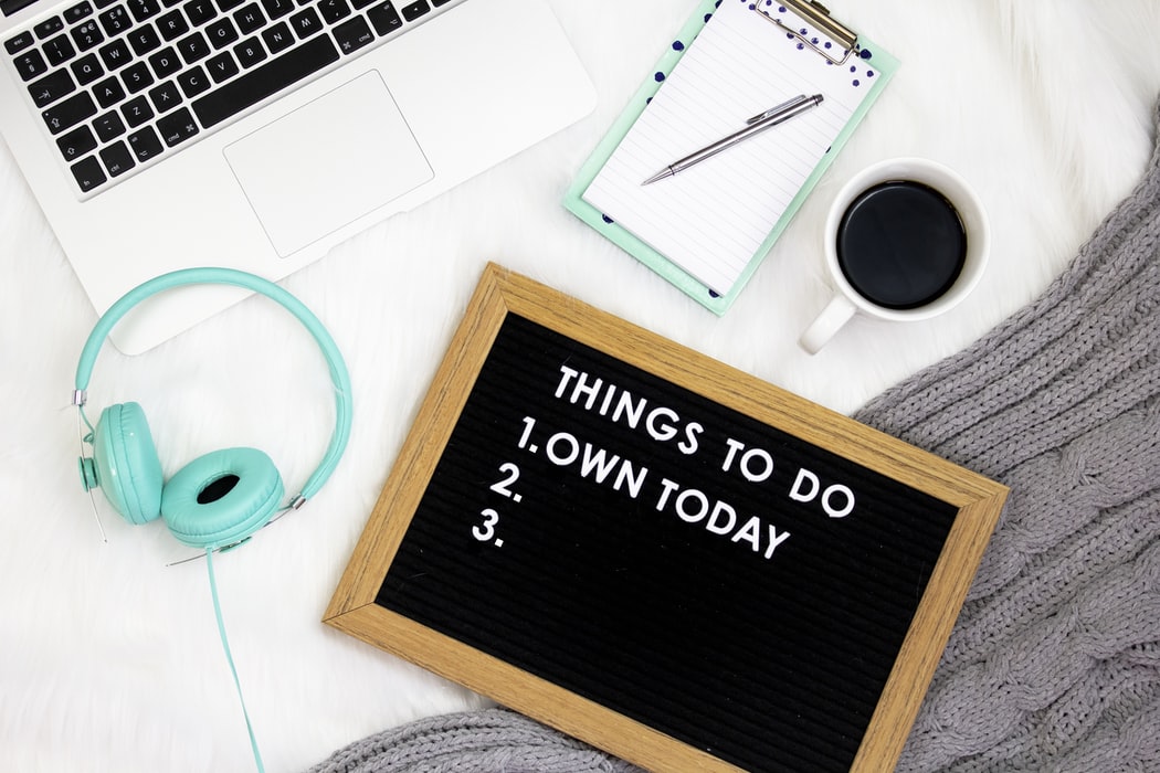 A sign surrounded by a computer, coffee, headphones, and a checklist that says “Things to do: 1. Own today 2. 3.”