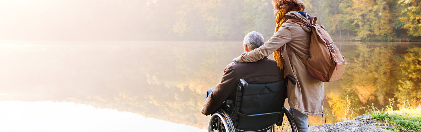 A woman with her hand over a man in a wheelchair looking out over a body of water