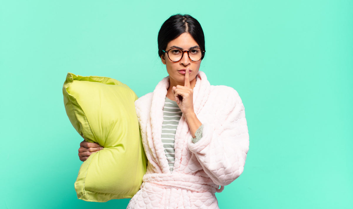 A woman holding a pillow in a robe with her index finger over her mouth