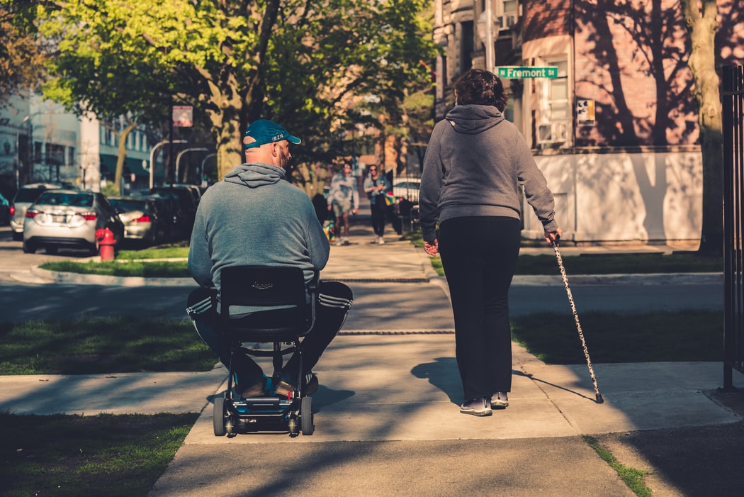 A man riding on a scooter and a woman using a cane to walk on a sidewalk