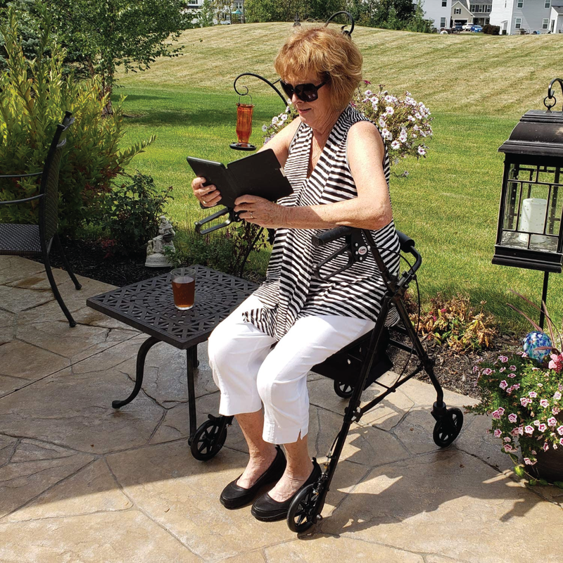 A woman sitting outside on the Carex Step ‘N Rest Rollator while reading a book