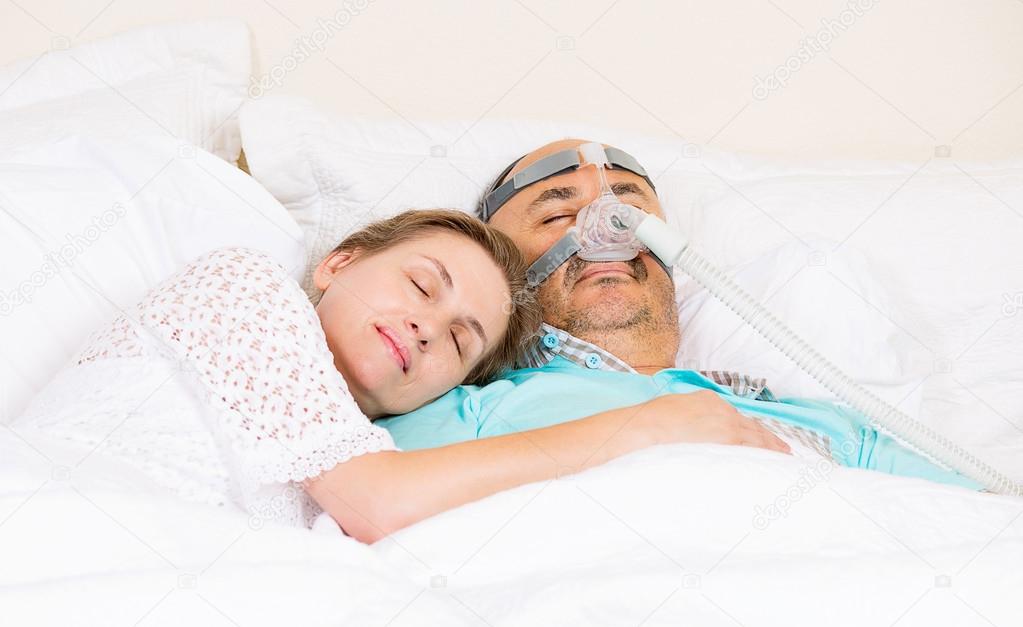 A man and woman sleeping in bed. The man has a CPAP mask on.
