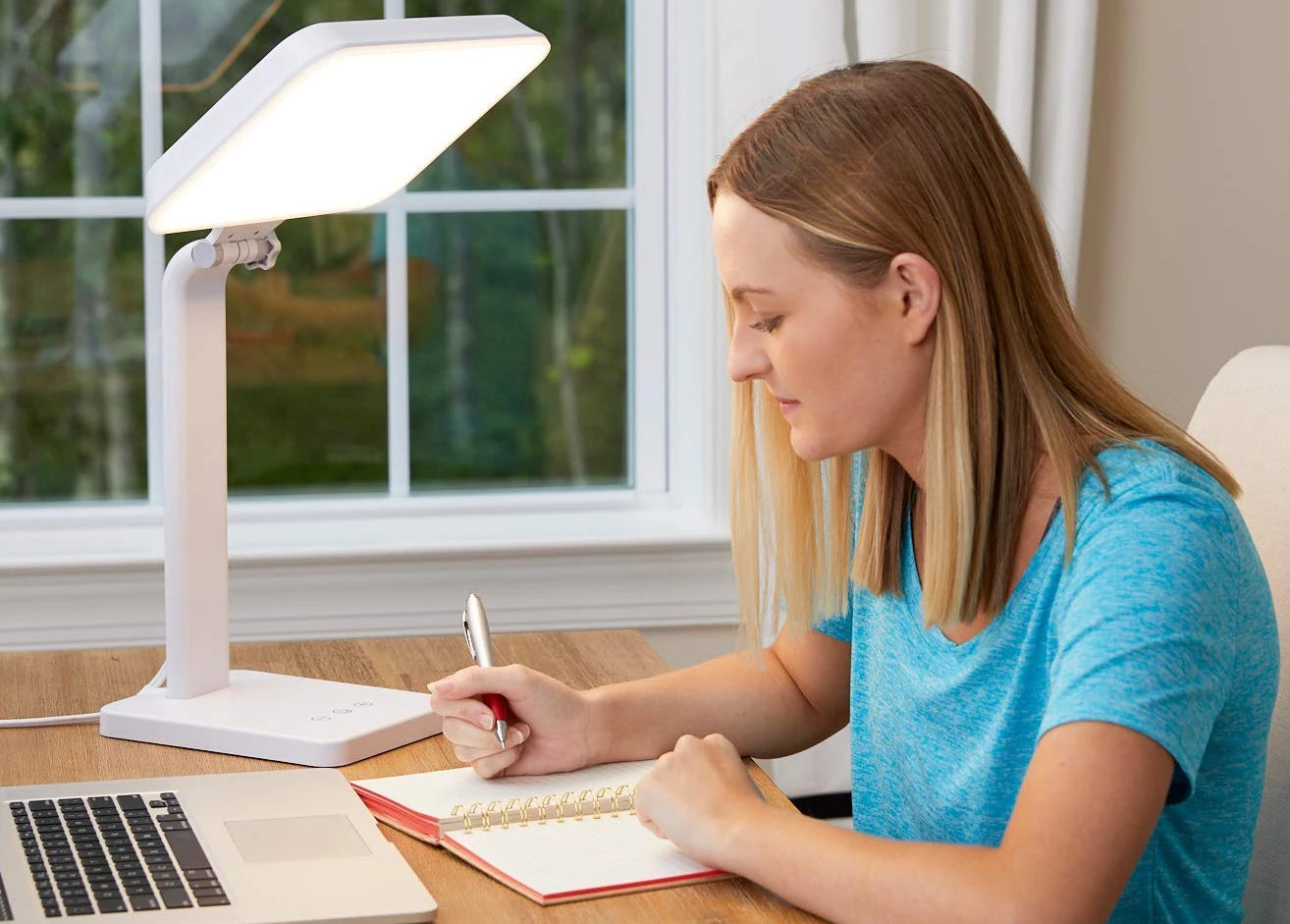 A woman sitting next to a light therapy lamp at her desk writing