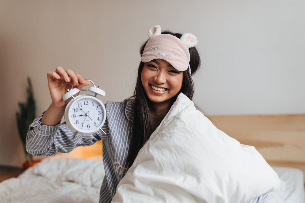 A smiling woman with a face mask on holding a clock in bed