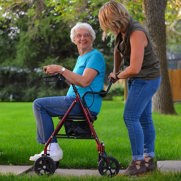 A caregiver on a sidewalk pushing an elderly woman sitting on the ProBasics Transport Rollator