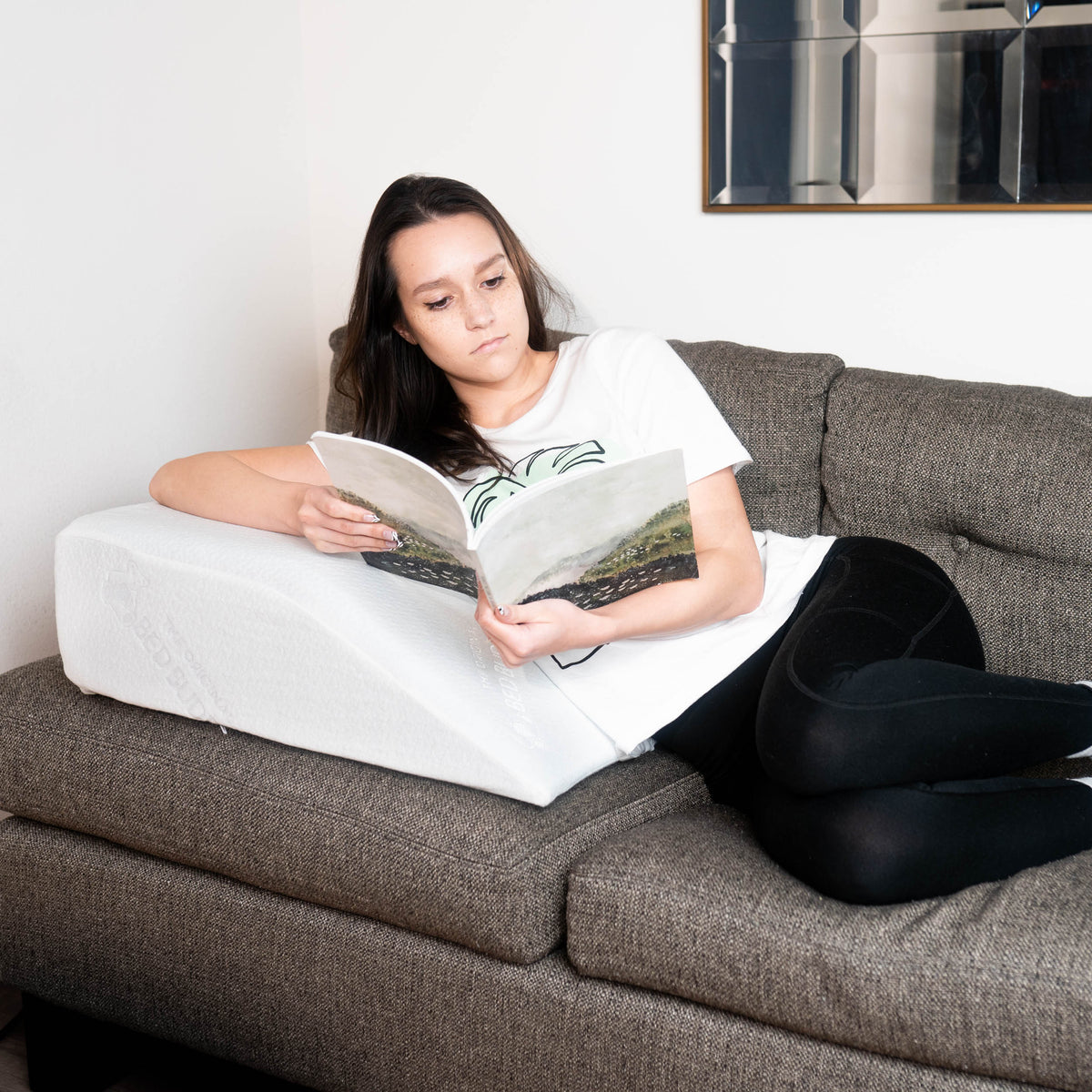 A woman reading a magazine on the couch while leaning on the Bed Buddy Leg Wedge Pillow
