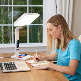 A woman writing next to a laptop in front of the TheraLite Aura Qi