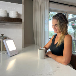 A woman enjoying a cup of coffee next to the TheraLite Glow 