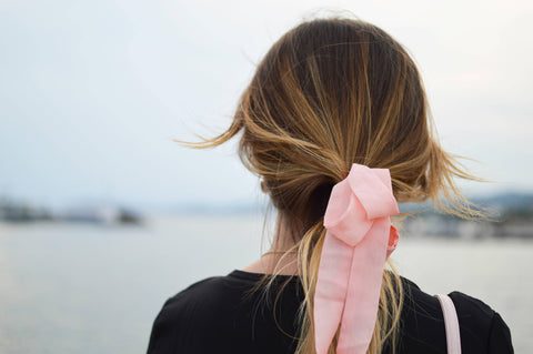 A young woman facing away from the camera with a light pink bow in her hair, which could be a fashion choice or a subtle, private political statement in support of breast cancer awareness.