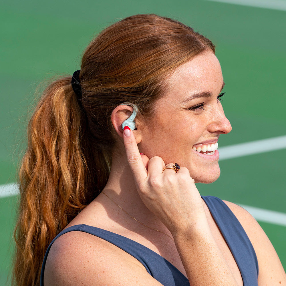 Frau, die GO Air Sport True Wireless Earbuds in Light Blue trägt und die Bedienelemente an der Seite des Ohrhörers berührt