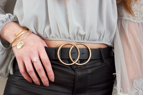 A close shot of a woman's midriff. She is wearing a grey cropped blouse and black leather pants with a thick, black belt that has two gold circles for the buckle. She has gold jewelry on the hand with its thumb hooked over her belt.