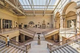 An interior shot of the Art Institute of Chicago. A large open set of stairwells in classical style. There are statues and other art pieces on the far walls of the upper balconies and in the center landing.