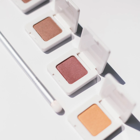 A set of three eyeshadows in white square compacts. Beside them is a silver handled makeup brush.