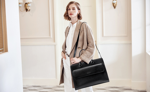 A woman in a long beige blazer and white button up shirt holds a black briefcase over one shoulder with the shoulder strap.