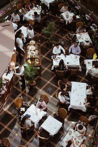 An overhead shot of a restaurant. Patrons sit at tables covered in white cloths in high-backed chairs. The waitstaff is in white shirts and black slacks.