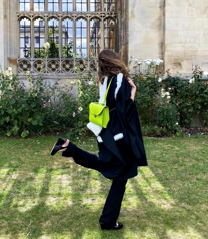 A woman dressed in black poses with her back to the camera, holding a bright yellow-green satchel bag.