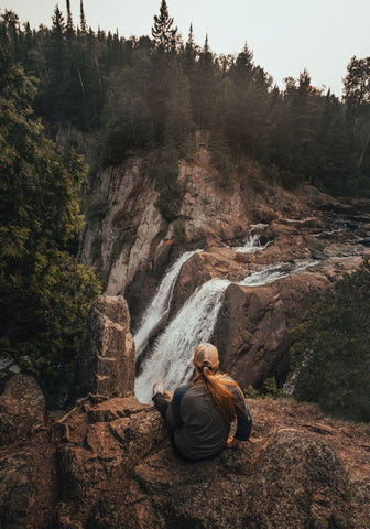 a woman sits with her back to the camera in front of a vista with a waterfall and a forest-covered mountain slope.