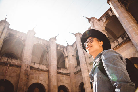 A man in a grey jacket, brimmed hat, and glasses stands above the camera, smiling. He is standing in what looks to be a stone castle courtyard at midday.