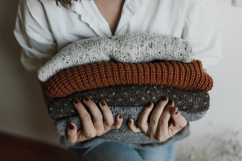 A close shot of a woman holding four thick knit sweaters. From top to bottom: a light speckled white, a dark orange, a dark grey with white dots, and a plain light grey.