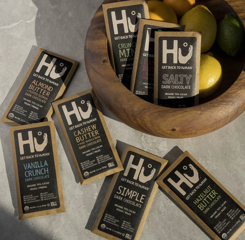 An assortment of Hu chocolate bars laid out on a marble counter and in a wooden bowl on top of some lemons. The bars include Almond Butter, Cashew Butter, Salty Dark Chocolate, and Simple Dark Chocolate.