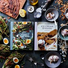 A flat lay of two recipe books surrounded by cooking ingredients and food including split hard boiled eggs, lemon, ice cream, and various herbs. The books are titled "Mighty Salads" and "Vegan Ice Cream and Friends."