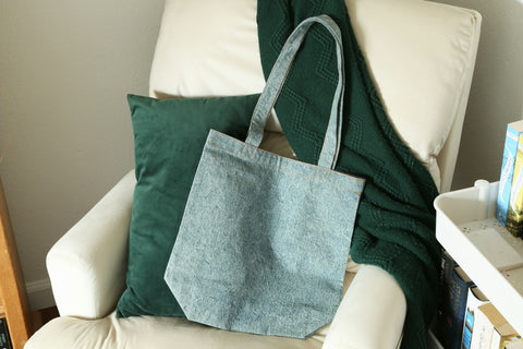 A light wash denim tote bag on a white chair with a green throw behind it.