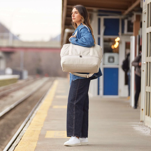 A woman in a denim jacket and black pants holds a white weekender tote.