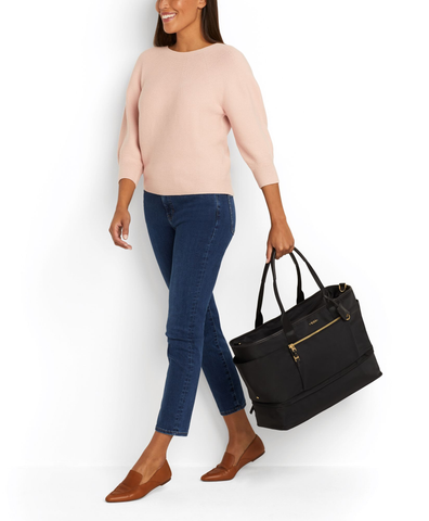 A woman in a light pink sweater and dark pants holds a black weekender tote.