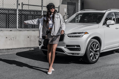 A woman stands in front of a white SUV. Her outfit is largely white, with a patterned skirt and lots of black accessories.