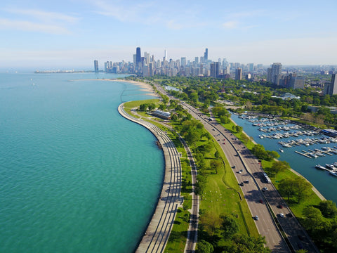 An overhead shot of the Lakefront. A highway runs parallel to the shore of a lake, which is cordoned off by green space and walking paths. In the distance is the skyline of Chicago.