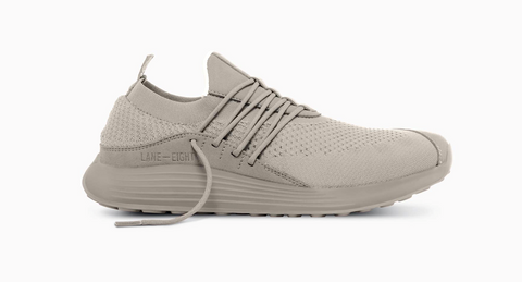 A tan-grey casual sneaker with laces across the middle. The upper appears to be made of mesh with leather accents.