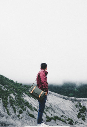 A man in a red windbreaker stands in front of a grey and green mountainside. He holds a green and tan messenger bag.