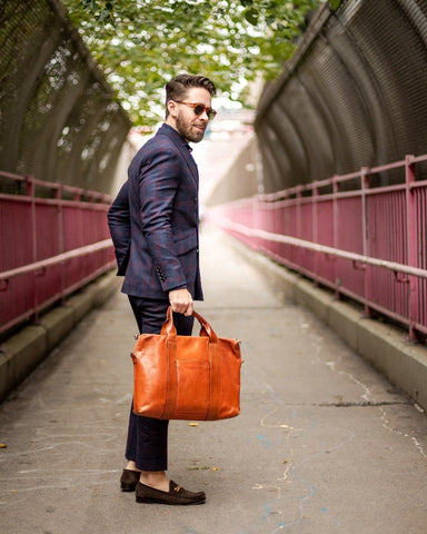 A man in a blue suit holds a light brown messenger bag as he poses on a concrete bridge.