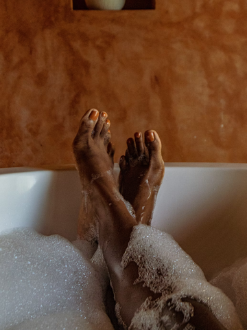A person's feet in a white bathtub. Their toenails are painted orange and their legs are covered in bubbles.